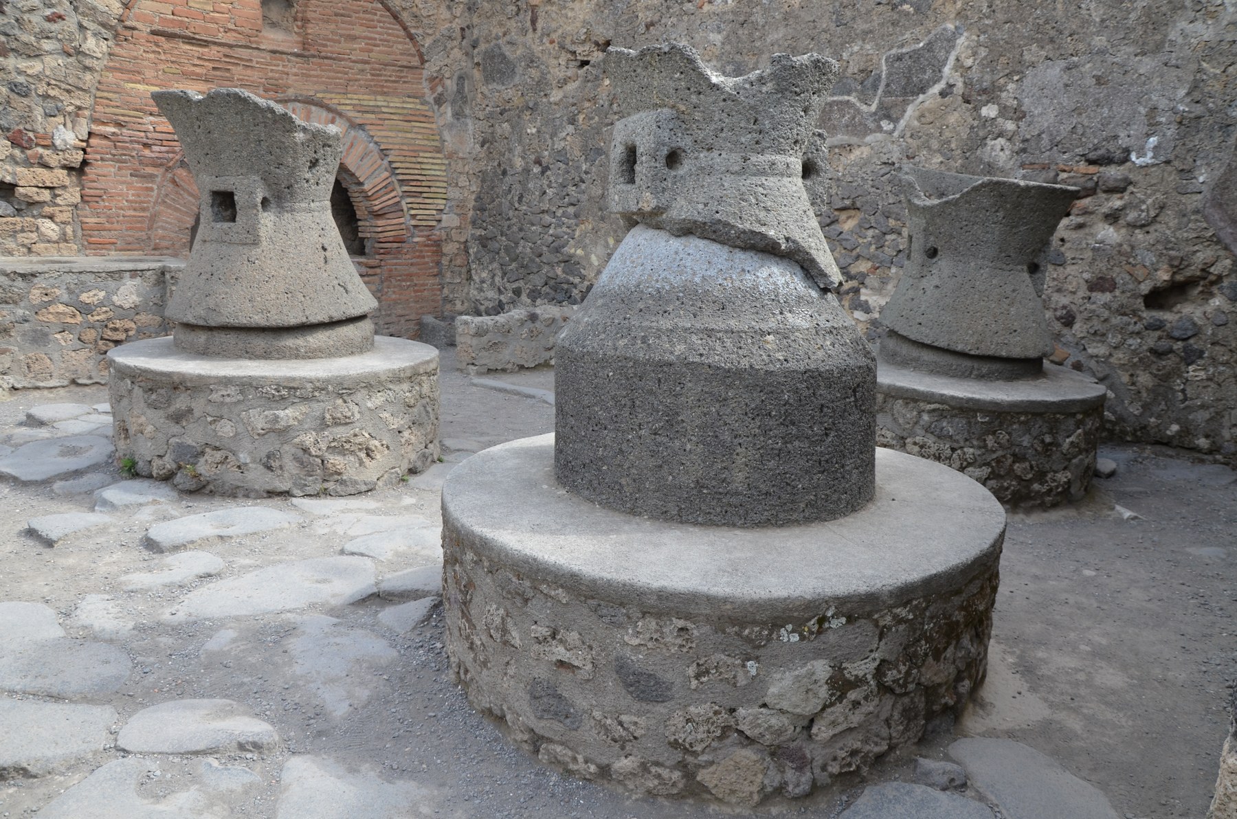 Large hourglass-shaped stone mills standing in a ruined Pompeian bakery.