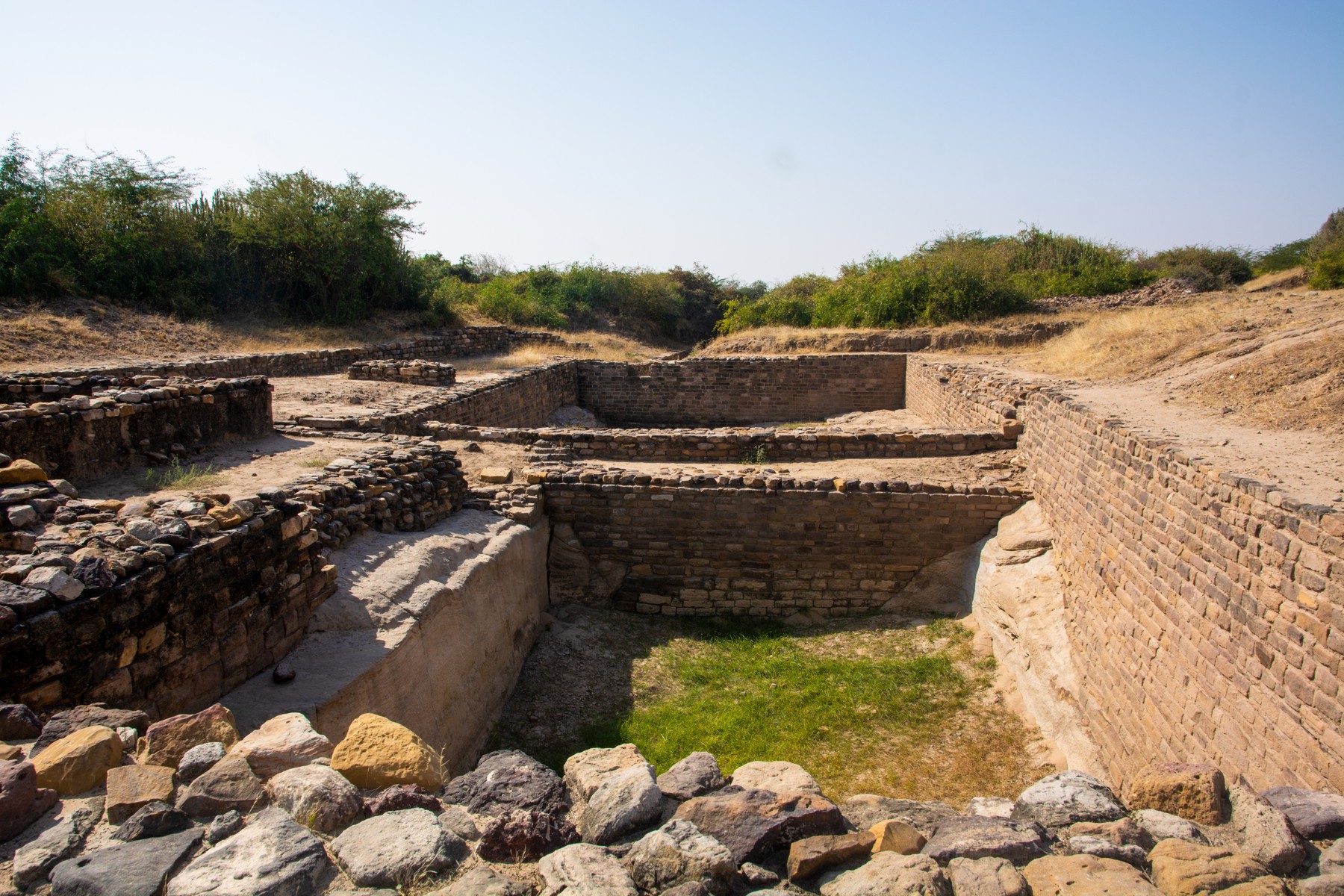 Excavated rectangular stone structures and sunken chambers at Dholavira, with vegetation beyond.