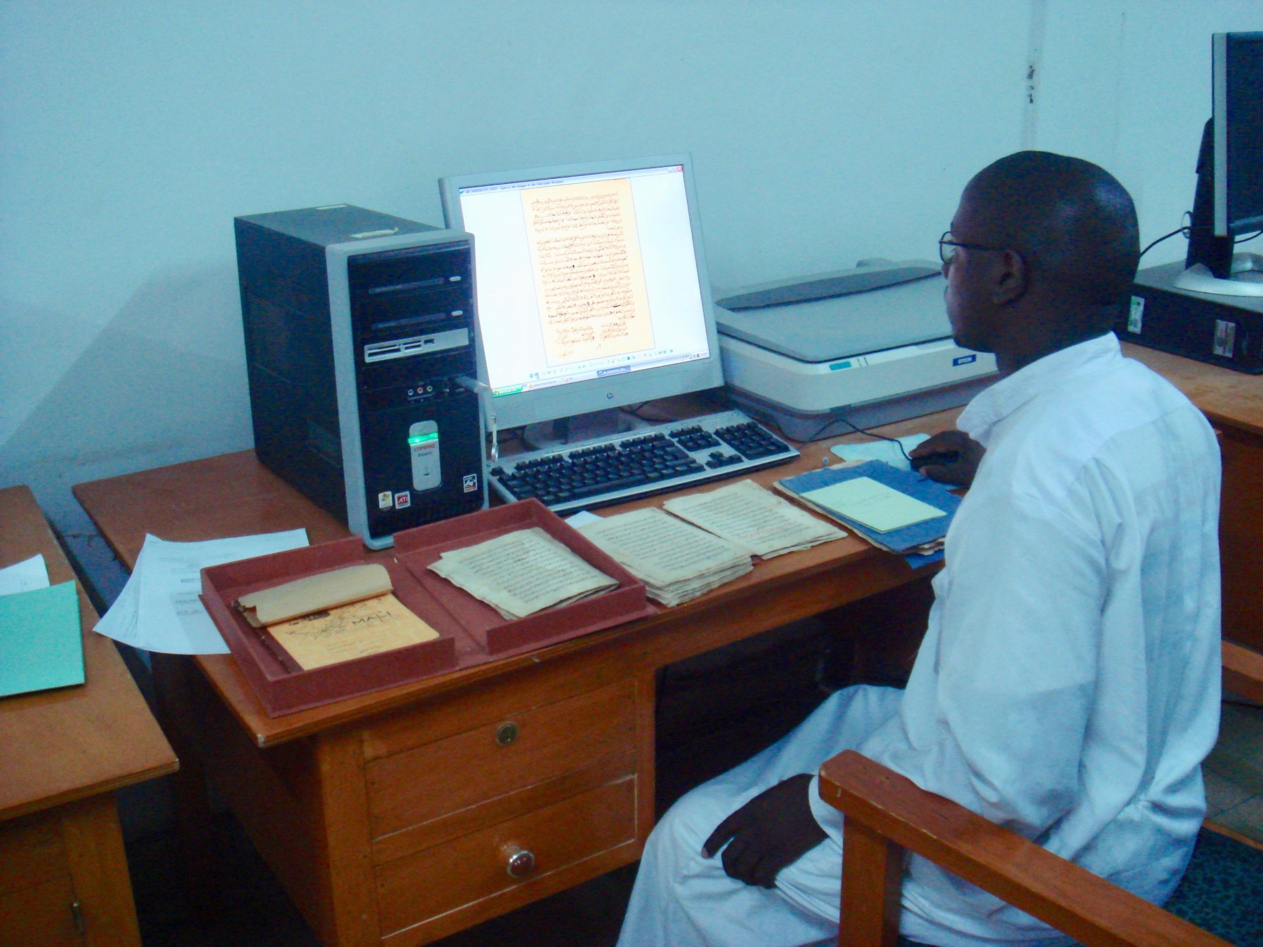 An archivist sits at a computer beside a scanner and open manuscript folders on a wooden desk.
