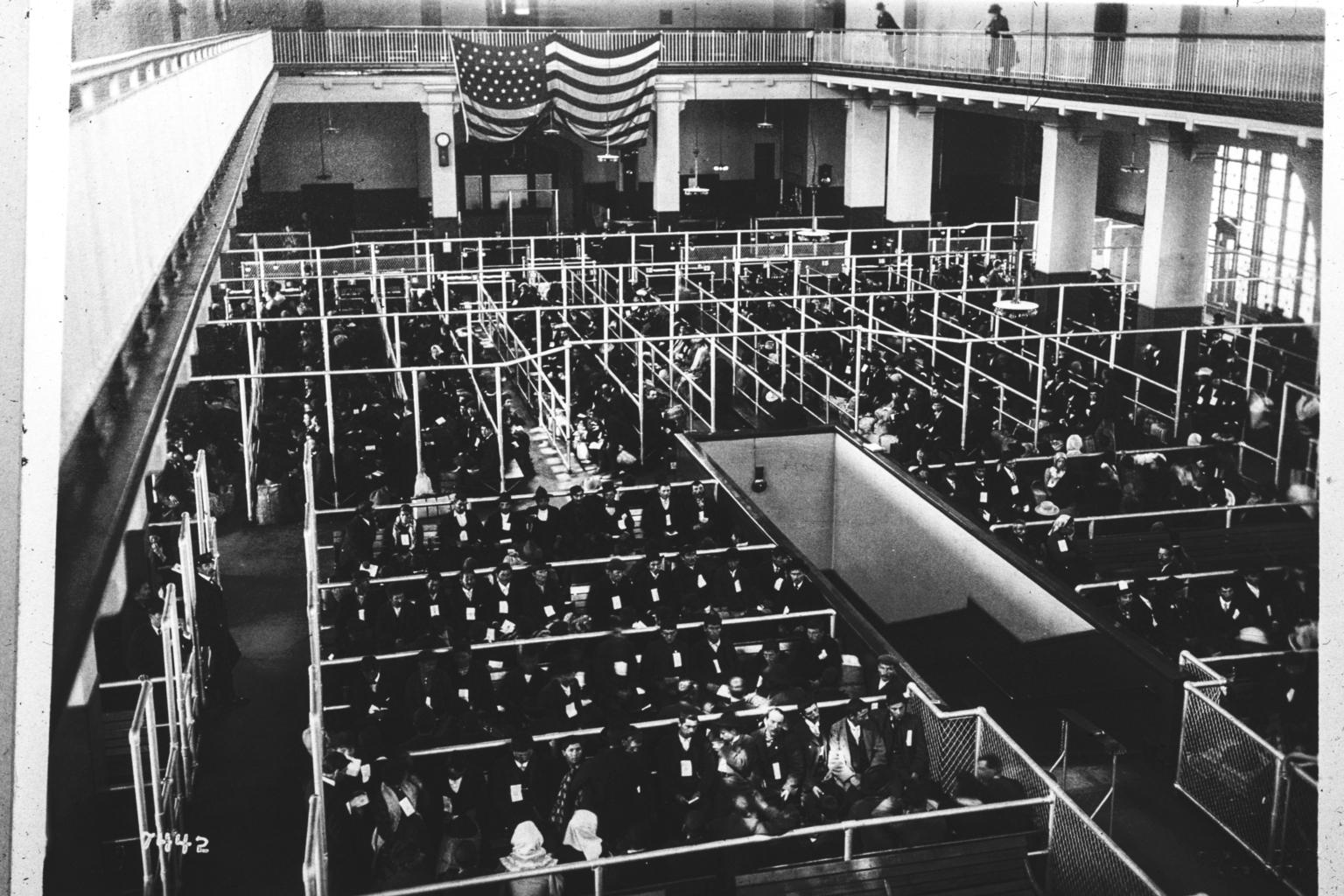 A high view across rows of crowded waiting enclosures beneath a large United States flag.