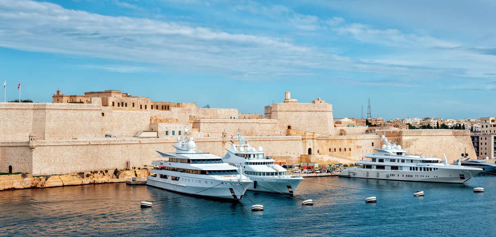 Panoramic modern view across water towards the stone walls of Fort St Angelo, with large yachts moored in front.