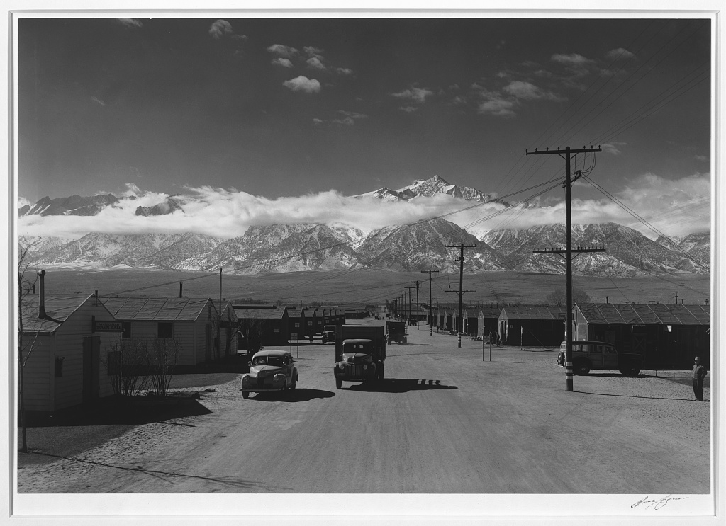 Cars and a truck on a broad Manzanar street between barracks, with utility poles and snow-covered mountains behind