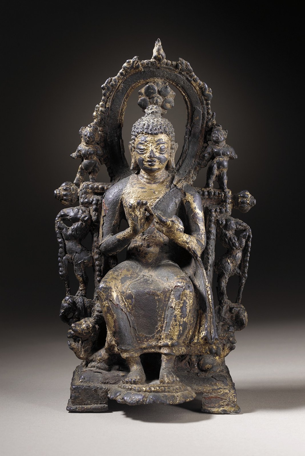 Small gilt copper-alloy Buddhist figure seated on an elaborate throne with hands raised before the chest.