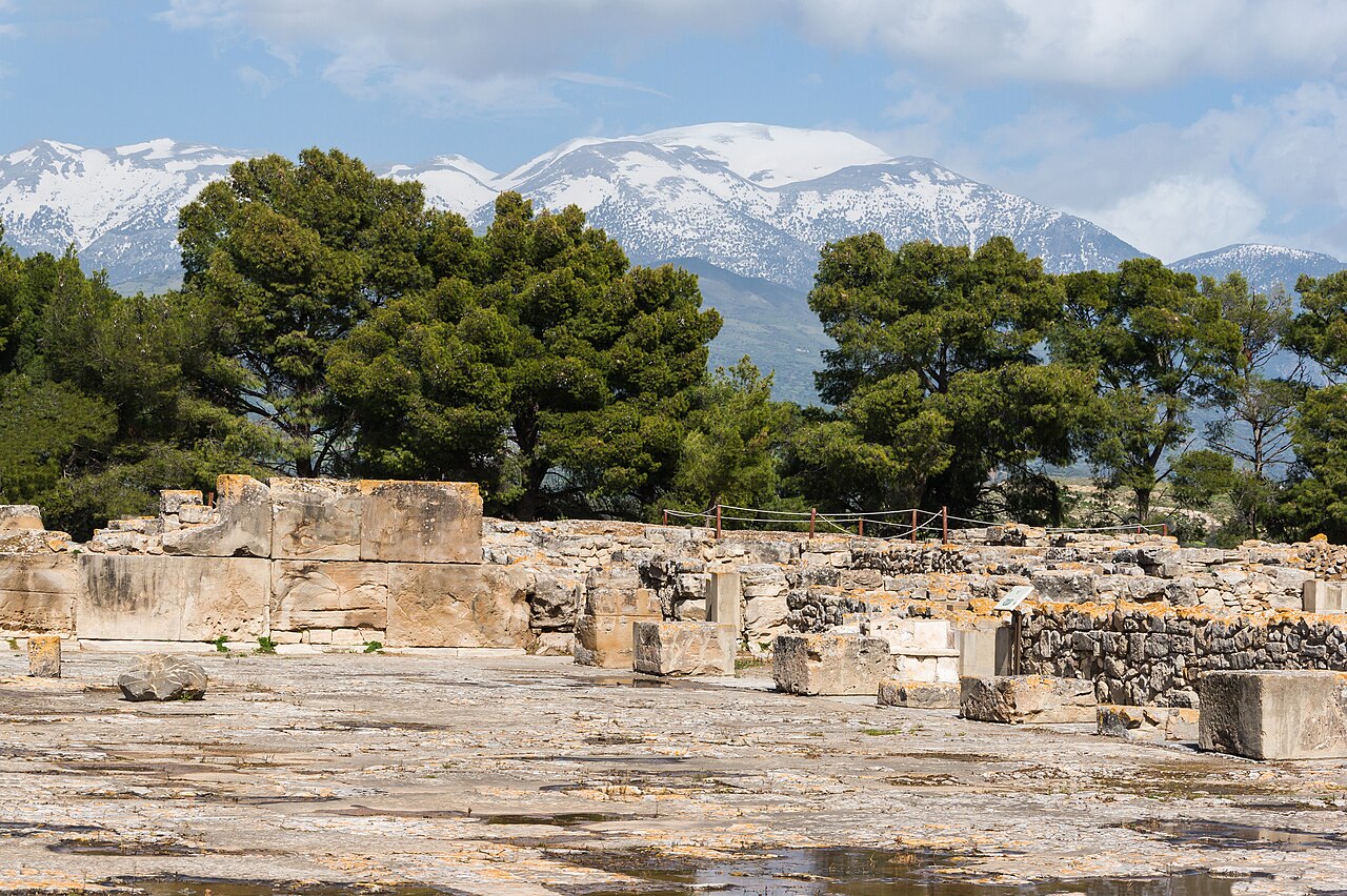 Stone paving and low ruined walls of the central courtyard at Phaistos, with trees and snow-covered mountains behind.