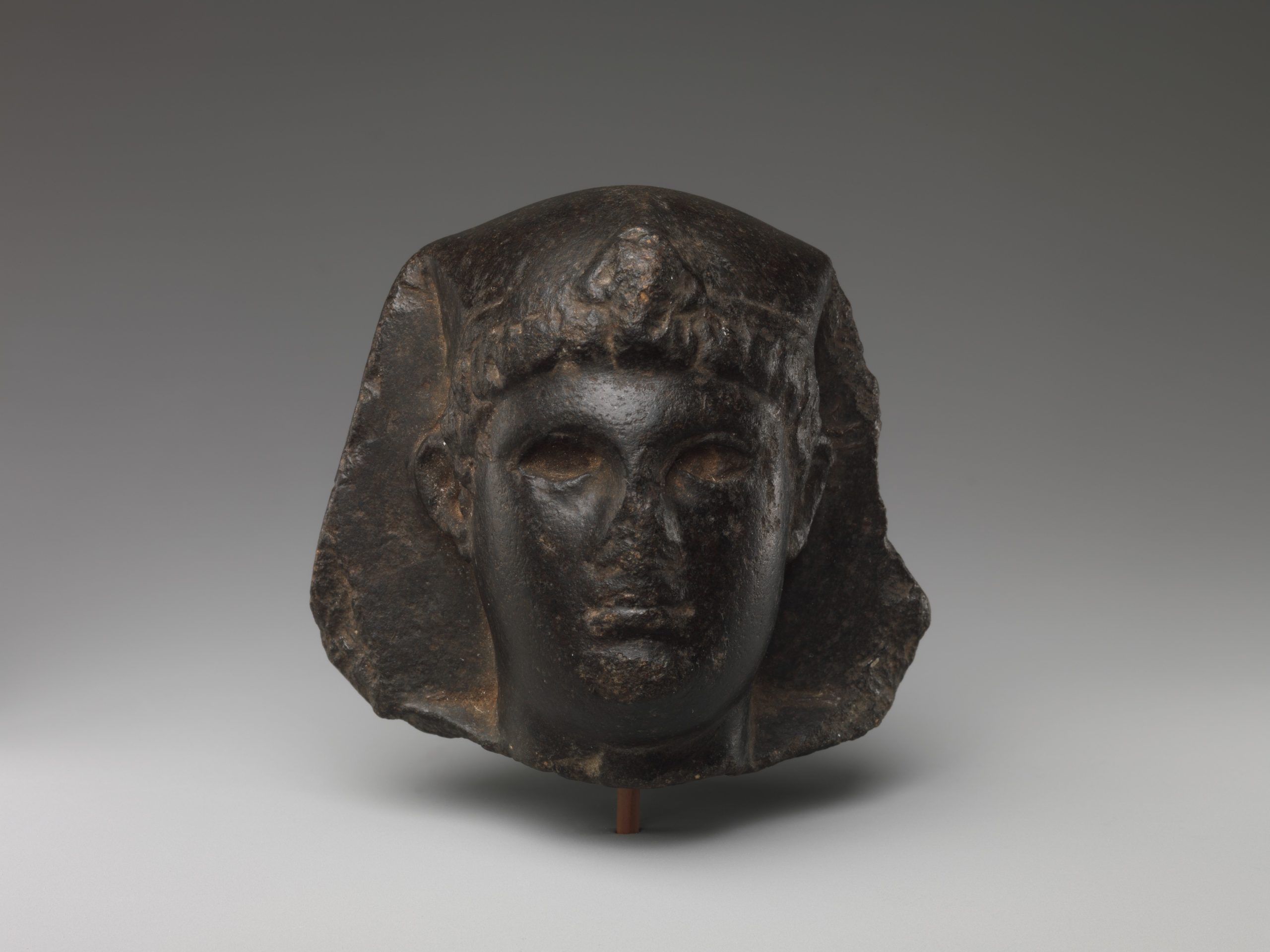 Dark stone head of a Ptolemaic ruler combining an Egyptian headdress with curled Greek-style hair.