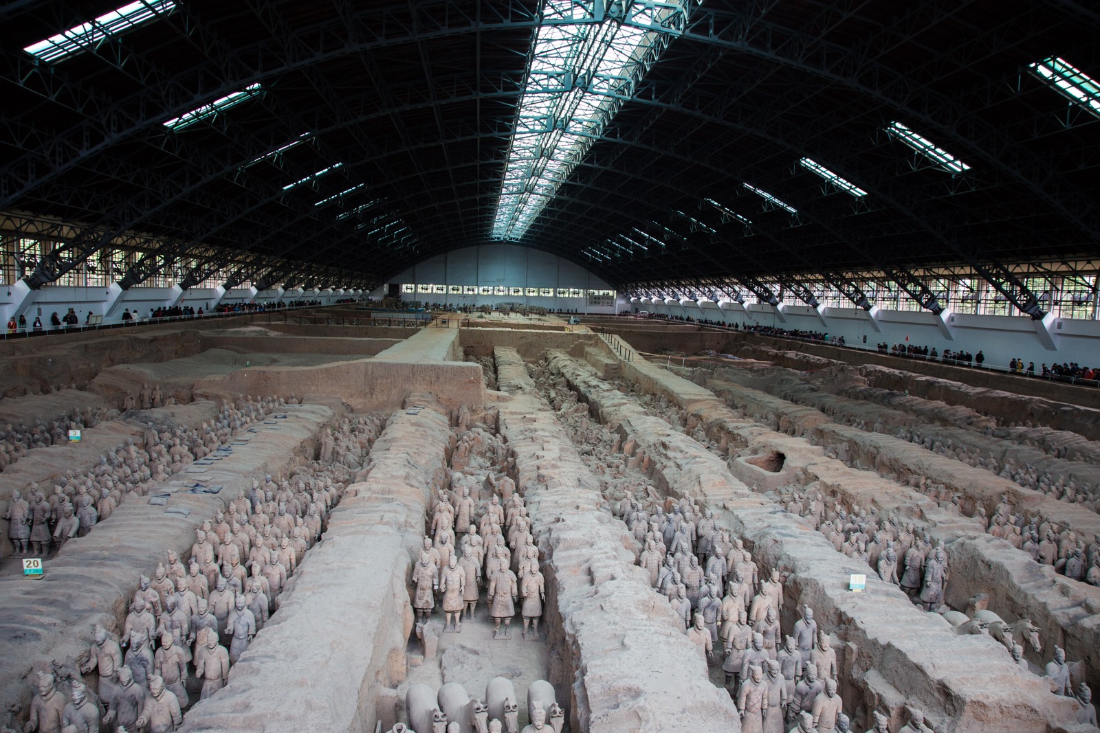 Rows of terracotta figures in long excavated corridors beneath a modern roof