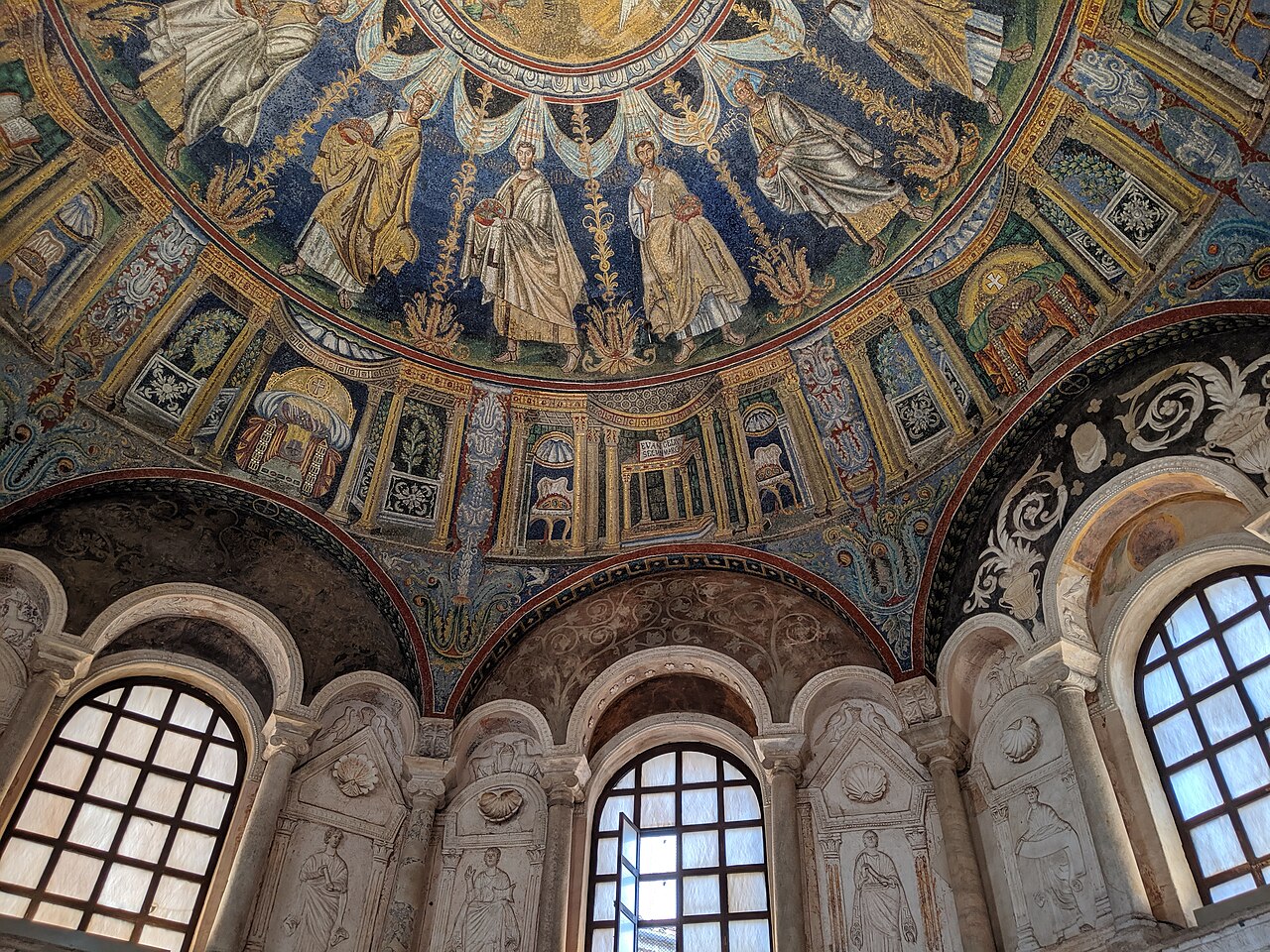 Curving Neonian Baptistery interior with apostles on a blue mosaic band above decorative thrones, stucco reliefs and arched windows.