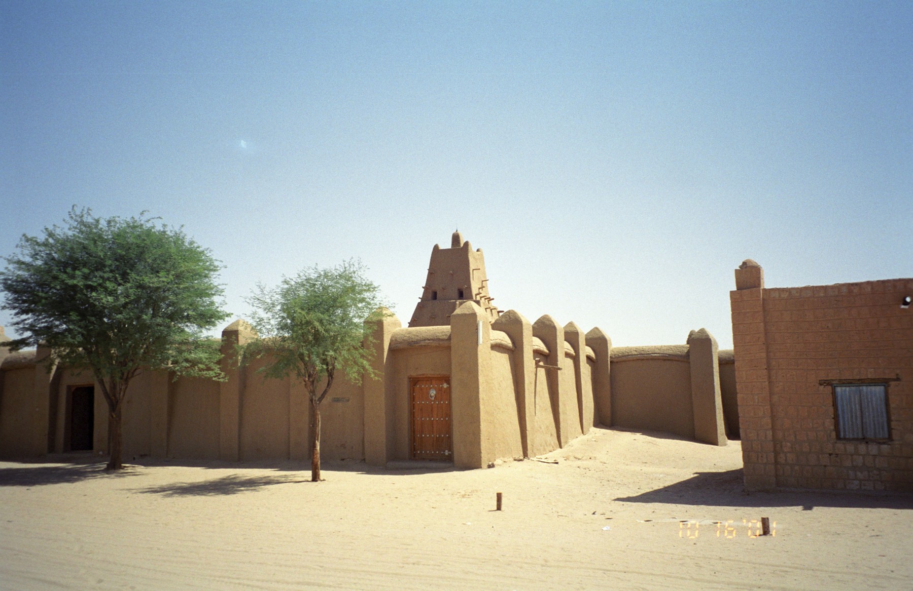 Sunlit earthen walls and projecting buttresses of Sankore Mosque, with trees beside an entrance.
