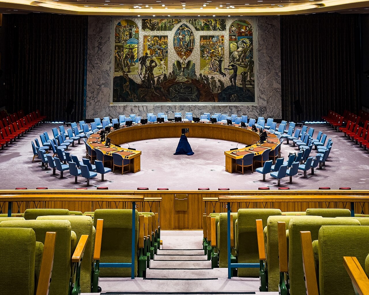 An empty United Nations Security Council chamber with a curved conference table, blue chairs and a large mural.