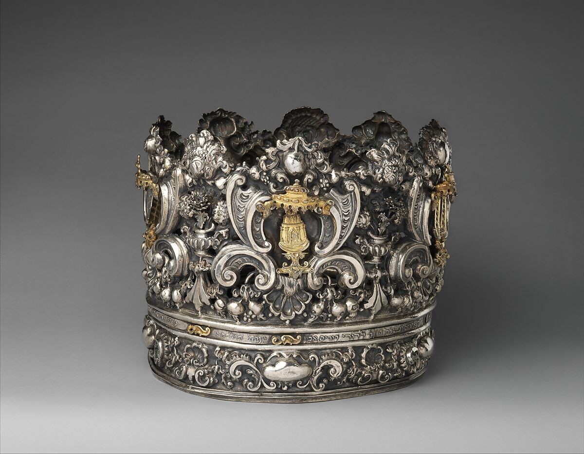 Ornate silver Torah crown with floral forms and ritual emblems against a gray background.